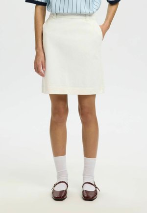 Žena v béžovej sukni typu A-line, bielych ponožkách a tmavočervených topánkach Mary Jane, stojaca s rukami v vreckách.