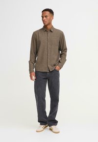Bruine casual overhemd met een borstzak, gecombineerd met donkergrijze jeans en beige sneakers met drie strepen. Glad materiaal met een subtiele textuur.