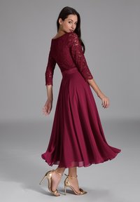 Robe bordeaux avec des manches en dentelle 3/4, jupe fluide et taille cintrée, associée à des sandales à talons dorées. Texture lisse et design élégant.