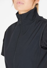 Gilet nere senza maniche con colletto alto, chiusura con zip, dettagli in tessuto testurizzato sulle spalle e materiale principale liscio. Tiretto della zip regolabile.