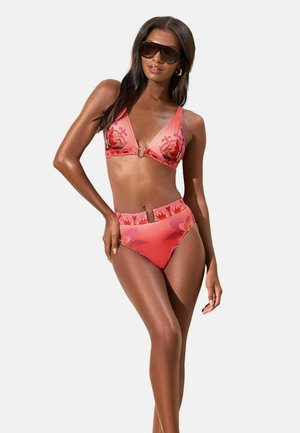 Kvinde iført koralrosa højtaljet bikini med blomsterprint og store mørke solbriller, poserer med den ene hånd på hoften mod hvid baggrund.