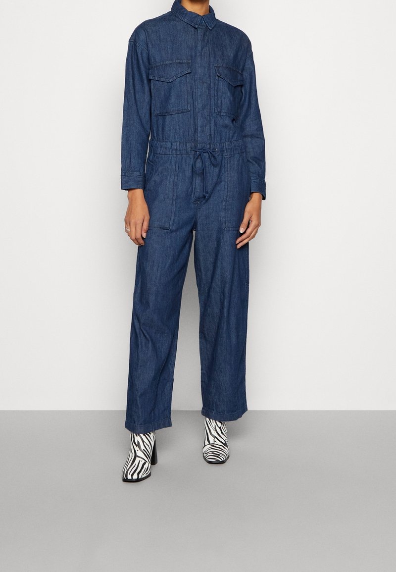Personne portant une combinaison en jean bleu à manches longues avec poches poitrine boutonnées, associée à des bottines imprimé zèbre noir et blanc.