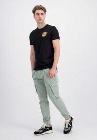 Čierne tričko s logom NASA, doplnené o svetlozelené cargo nohavice s viacerými vreckami. Outfit dopĺňajú čierne a šedé tenisky.