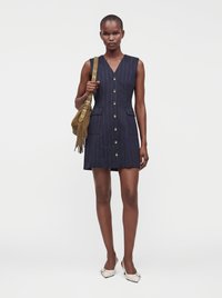 Femme portant une robe sans manches bleu marine à rayures fines avec des boutons, tenant un sac à épaule marron à franges et portant des chaussures blanches à bout pointu.