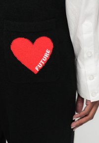 Černý svetr s červenou pletenou nášivkou srdce a bílým nápisem "FUTURE" na dolním levém kapse; hladká textura a ležérní design.