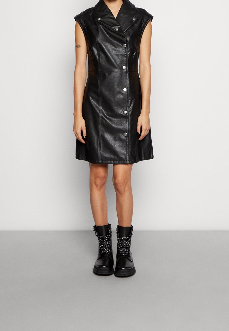 Robe noire en cuir sans manches boutonnée, portée avec des bottines noires à lacets sur une personne debout devant un mur uni.