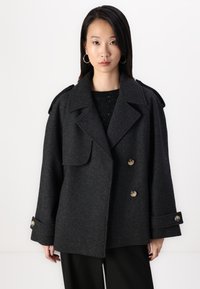 Manteau double boutonnage en mélange de laine grise avec grands revers, poches avant et boutons en laiton. Le design comprend des épaulettes et des manches larges.