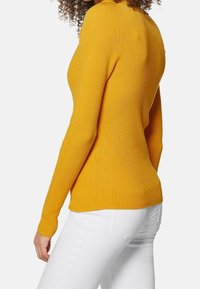 Femme aux cheveux bouclés portant un pull côtelé moutarde ajusté et un pantalon blanc, vue de profil et de dos.