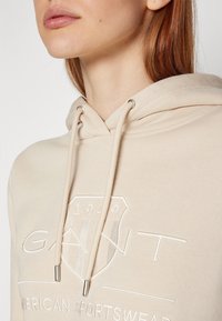 Gros plan d'une personne portant un sweat à capuche beige avec « GANT 1949 American Sportswear » brodé sur le devant, montrant le cou et le bas du visage.