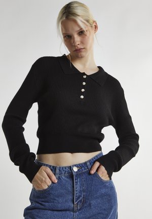 Jonge vrouw met blond haar die een zwarte geribbelde cropped trui met knopen en hoog getailleerde blauwe spijkerbroek draagt, handen in de zakken.
