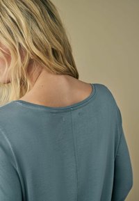 Helehelesinine lühikeste varrukatega topp, millel on lai kaelus ja vabam sobivus. Pehme tekstuur, millel on seljal õmbluse detail.