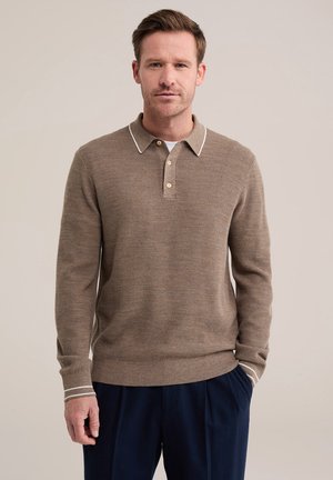 Homme portant un polo marron en maille à manches longues avec bordure blanche et un pantalon bleu foncé, debout devant un fond uni.