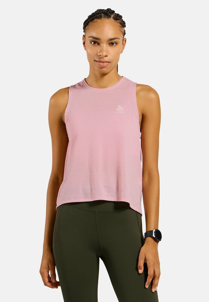 Junge Frau mit geflochtenem Haar, die ein ärmelloses pinkes Sporttop, olivgrüne Leggings und eine schwarze Smartwatch trägt und vor einem einfarbigen Hintergrund steht.