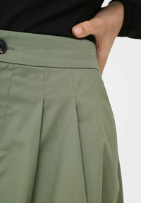 Jupe verte plissée avec fermeture à boutons à la taille. Fabriquée en tissu texturé, elle présente des lignes lisses et une coupe ajustée.