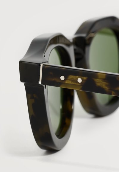 Lunettes de soleil rondes sombres en écaille avec des montures épaisses et des verres teintés verts, montrant la charnière et deux rivets argentés sur la branche.