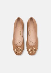 Paire de ballerines en cuir beige avec boucles décoratives rondes en or et semelles intérieures perforées et rembourrées sur fond blanc.