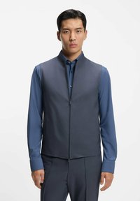 Gilet gris foncé avec col montant et fermeture éclair à l'avant, associé à une chemise bleue à manches longues. Caractérisé par des poches latérales et un design épuré.