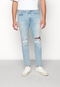 Ljust blå denimjeans med slitna detaljer, med revor vid knäna och en slim fit-design. Matchas med vita sneakers.