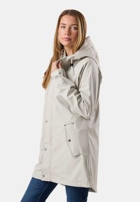 Femme tenant le col d'un imperméable à capuche beige clair avec boutons-pression et poches à rabat inclinées, portant un jean bleu.