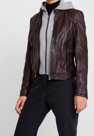 Osoba ubrana w ciemnobrązową skórzaną kurtkę na szarej bluzie rozpinanej na zamek i czarnych spodniach, ręce swobodnie opuszczone wzdłuż ciała, na jednolitym tle.