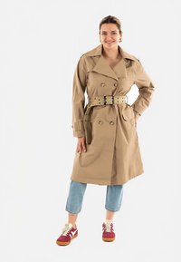 Gabardina beige con diseño de doble abotonadura, cinturón en la cintura y solapas grandes. Combinada con vaqueros cortos azul claro y zapatillas rojas.