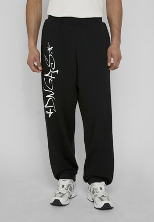 Black loose-fit sweatpants with white graffiti-style text on left leg, worn with white sneakers and a white shirt.