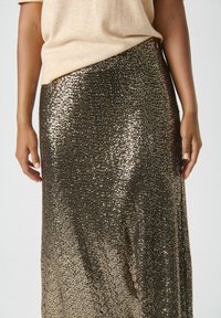 Gonna di paillettes con un motivo luccicante in oro e nero, caratterizzata da una forma dritta e una texture liscia. Indossata con un top beige.