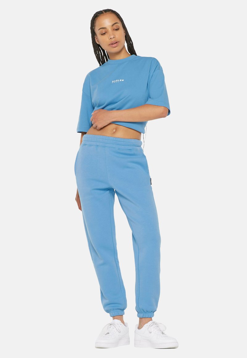 Gekürztes blaues Sweatshirt mit dem Schriftzug "HARLEM", kombiniert mit passenden blauen Jogginghosen. Beide sind aus weichem Stoff mit elastischen Bündchen gefertigt. Weiße Sneaker.