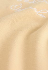 Vaaleankeltainen kangas, jossa on sileä rakenne ja valkoista brodeerausta. Materiaali näyttää pehmeältä ja hieman venytetyltä.
