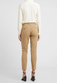 Hnědé kalhoty na míru se slim fit, dvěma zadními kapsami a hladkým povrchem, v kombinaci s světlou košilí s dlouhým rukávem a sandály na podpatku.