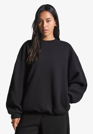 Young woman with long dark hair wearing a loose black sweatshirt and black pants, standing against a white background, looking directly at the viewer.