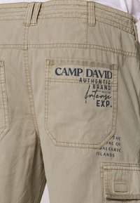 Beige cargo šortky s textúrovanou tkaninou, dvoma zadnými vreckami a potlačeným logom na jednom vrecku. Obsahujú zapínanie na gombík a slučku.