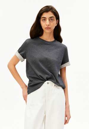 Vrouw met lang donker haar draagt een donkergrijs T-shirt met opgerolde mouwen, ingestopt in een hoge witte broek, tegen een effen achtergrond.