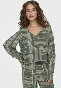 Top verde oliva con scollo a V e maniche lunghe, caratterizzato da un motivo astratto a strisce bianche. Tessuto morbido con una vestibilità comoda.