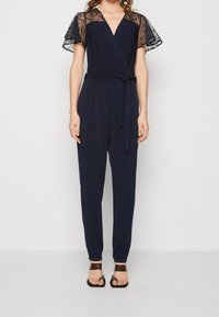 Combinaison bleu marine avec manches courtes en dentelle, taille ceinturée et jambes fuselées. Tissu lisse avec un léger éclat et décolleté en V profond.