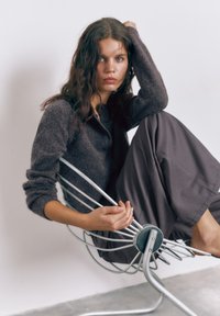 Ung kvinde med langt hår sidder barfodet i en moderne metalstol iført en mørk sweater og løse bukser foran en ensfarvet baggrund.