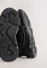 Paire de chaussures de sport noires debout montrant des semelles en caoutchouc texturées détaillées sur un fond clair uni.