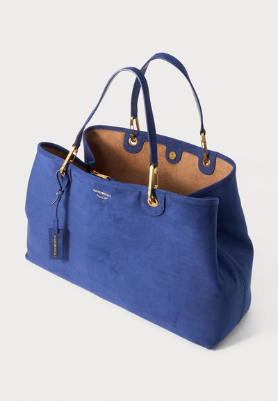 Sac fourre-tout bleu en daim avec quincaillerie dorée, deux poignées et un intérieur en liège. Comprend une étiquette et une poche zippée à l'intérieur. Texture lisse.