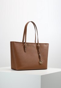 Sac fourre-tout en cuir marron avec une forme structurée, des poignées doubles et un charm logo doré. Dispose d'une surface texturée et d'un branding subtil.