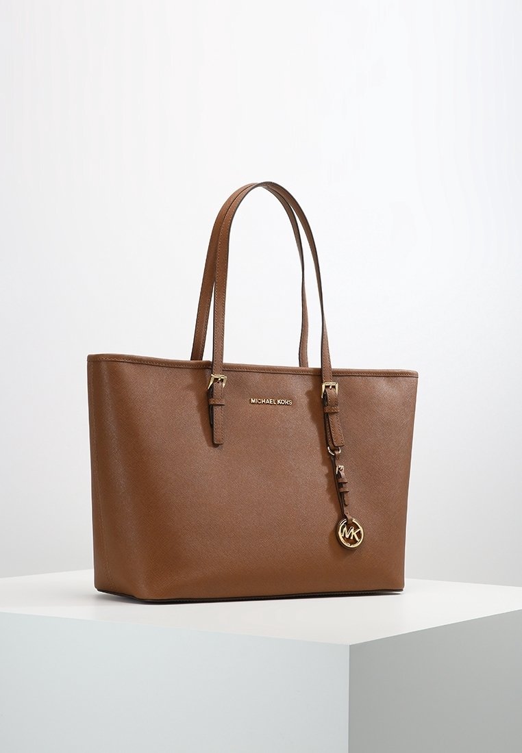 Sac fourre-tout en cuir marron avec une forme structurée, des poignées doubles et un charm logo doré. Dispose d'une surface texturée et d'un branding subtil.
