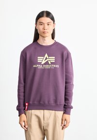 Purple sweatshirt with ribbed collar and cuffs, featuring a yellow logo and text “ALPHA INDUSTRIES” on the front. Red tag on the hem.