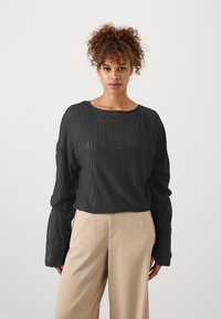 Haut noir à manches longues, côtelé et à coupe décontractée, avec un col bateau, fabriqué dans un tissu léger, texturé avec un effet légèrement transparent. Assorti à un pantalon beige.