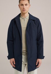 Chubasquero azul marino con un cuello estructurado, cierre de botones y un suéter beige texturizado debajo. El modelo lleva pantalones de color claro.