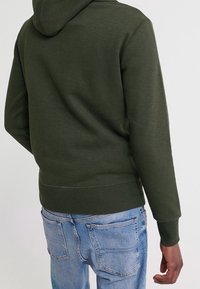 Felpa verde scuro con una finitura testurizzata, dotata di cappuccio con cordino e vita a strisciata. Abbinata a jeans chiari con effetto usurato.