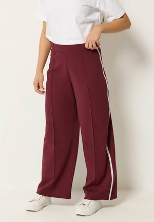 Kvinde iført bordeaux vidde bukser med hvide sidelister, kombineret med hvide sneakers og en hvid kortærmet top.