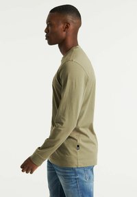 T-shirt a maniche lunghe verde oliva in cotone, con scollo rotondo, etichetta sul lato e vestibilità rilassata, abbinato a jeans blu.
