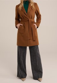 Bruine jas met ceintuur en reverskraag, voorzien van zakken, gedragen over een groene coltrui en grijze, breed uitlopende broeken met patroon.