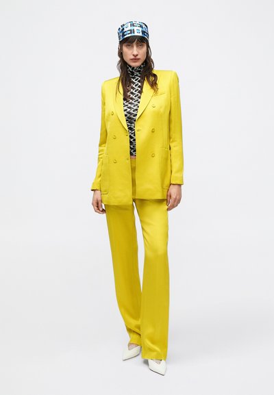 Abito giallo su misura con giacca doppiopetto e pantaloni a gamba larga, abbinato a un top nero e bianco con motivo e a un foulard blu con fantasia.