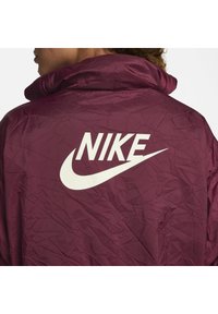 Bordo jakna z visokim ovratnikom, ki ima na zadnji strani velik bel Nike logo. Material ima nagubano teksturo.