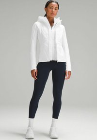 Veste blanche légère avec capuche, texture matelassée, fermeture éclair à l'avant et manches longues ; associée à des leggings bleu marine et des baskets blanches.